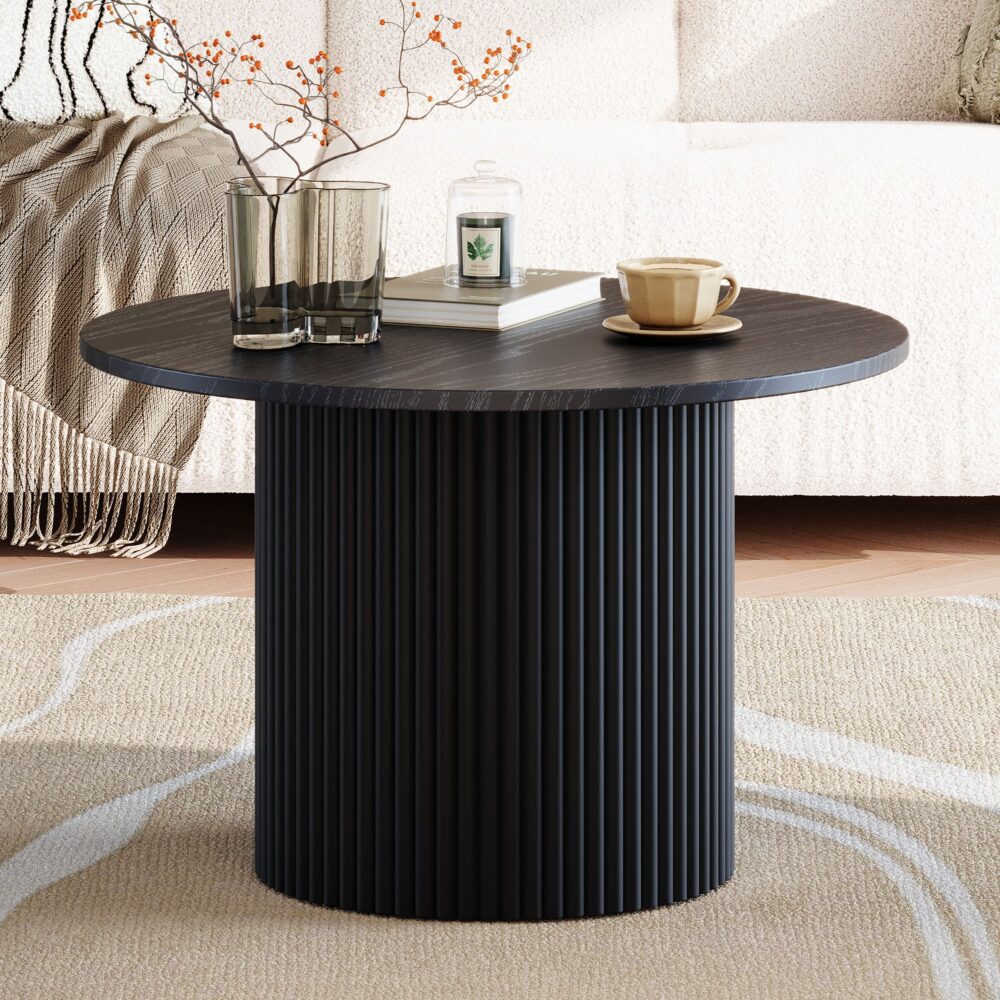 Black round coffee table with fluted cylindrical pedestal base on cream rug, books glasses and coffee cup on top