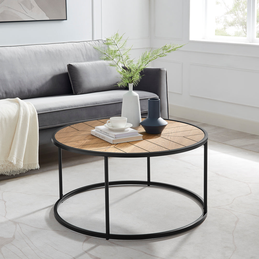 Round coffee table with sunburst wood top and black metal frame in living room with grey sofa, vases and cup on top