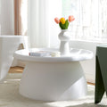 Round white coffee table with tray top styled with a vase of tulips, a book and a candle ornament near a window in a bright