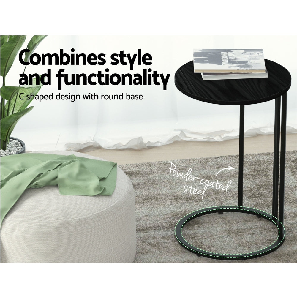 Round black C-shaped side table with powder-coated steel base callout, magazine on top, pouf and plant beside, living room