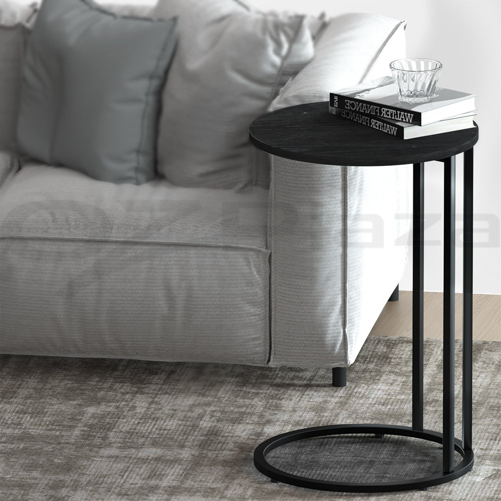 Round black side table with wood-grain top and black C-shaped metal frame with circular base, books on top, beside grey sofa