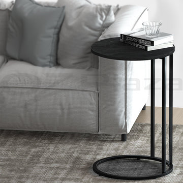 Round black side table with wood-grain top and black C-shaped metal frame with circular base, books on top, beside grey sofa