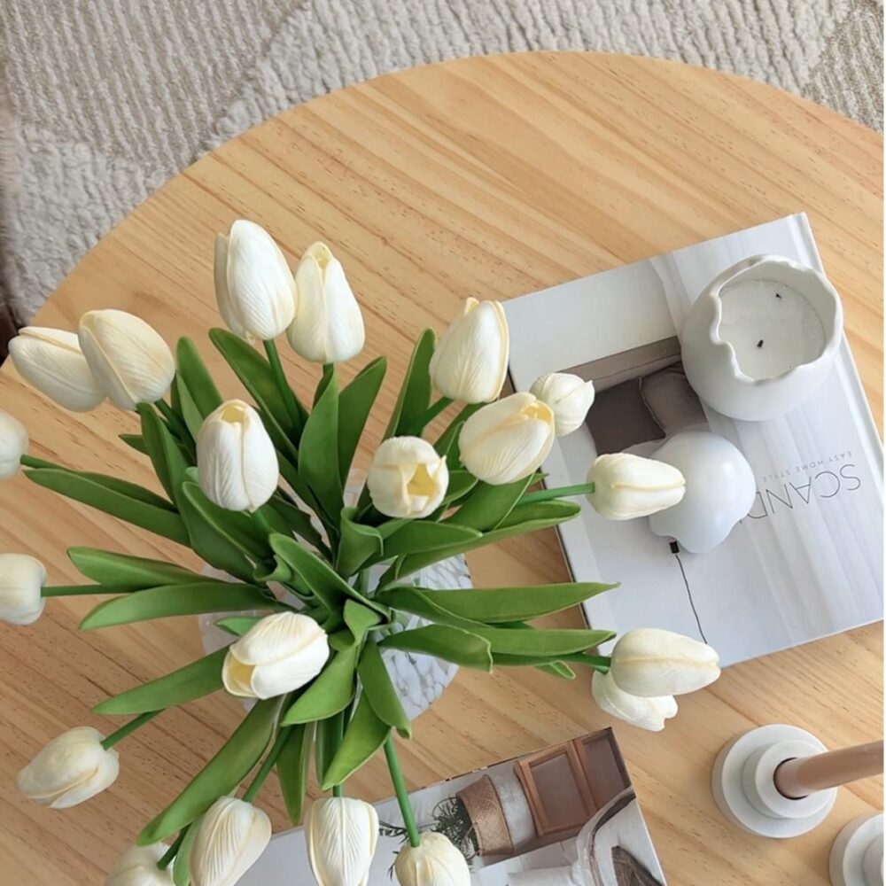 Overhead view of round natural pine coffee table top with white tulips in glass vase, open magazine and white ceramic candle