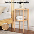 Round two-tier natural wood side table with twisted metal bar spindles, styled beside a bed with books on lower shelf and