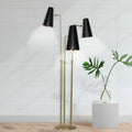 Three-arm floor lamp lit against white panelled wall beside glass vase with tropical leaves on white pedestal