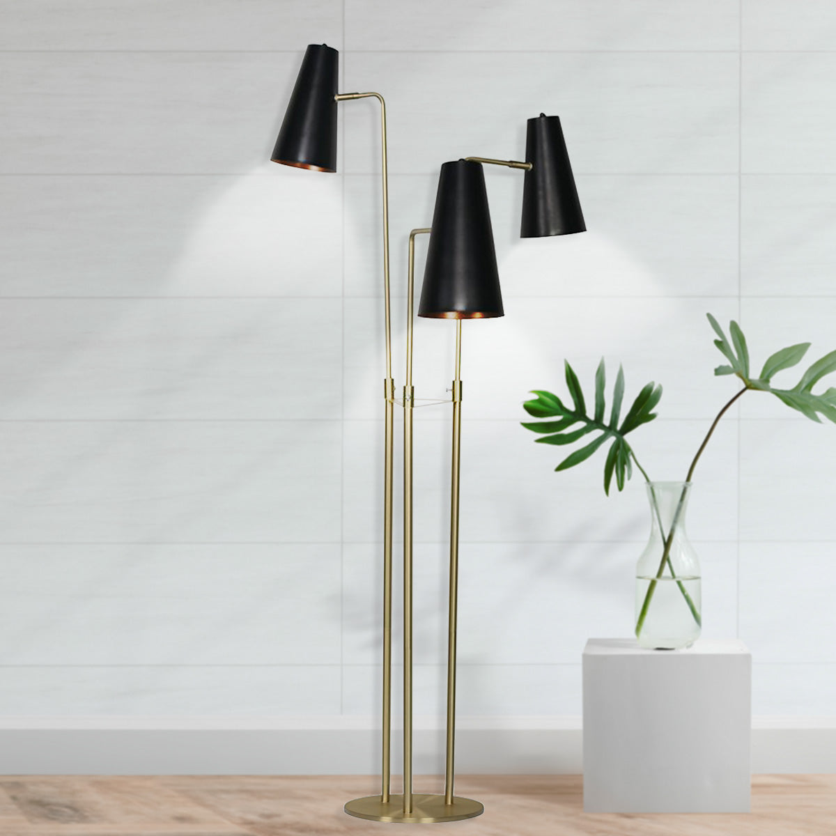 Three-arm floor lamp lit against white panelled wall beside glass vase with tropical leaves on white pedestal