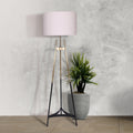 Gradient tripod floor lamp with white drum shade beside tropical plant in grey pot against concrete wall