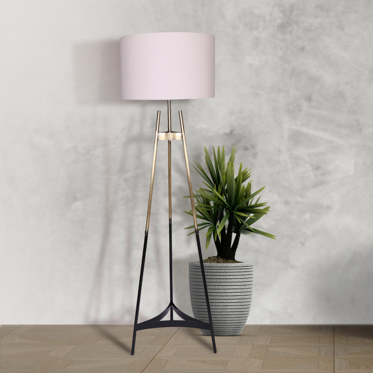 Gradient tripod floor lamp with white drum shade beside tropical plant in grey pot against concrete wall