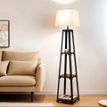 Tapered black shelf floor lamp with beige fabric shade lit in living room scene next to beige sofa with wall art