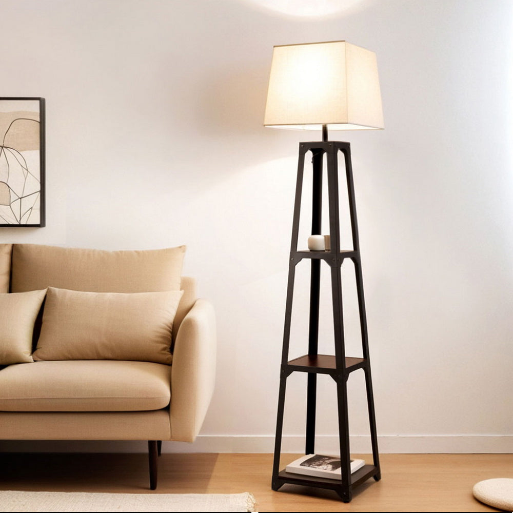 Tapered black shelf floor lamp with beige fabric shade lit in living room scene next to beige sofa with wall art