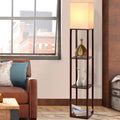 Walnut shelf floor lamp with lit white square shade in living room beside brown leather sofa, shelves holding decor and books