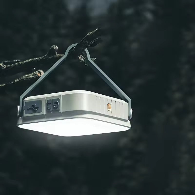 Square LED camping lantern hanging from tree branch by hook, USB port and buttons on side, dark forest background