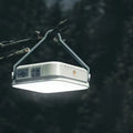 Square LED camping lantern hanging from tree branch by hook, USB port and buttons on side, dark forest background