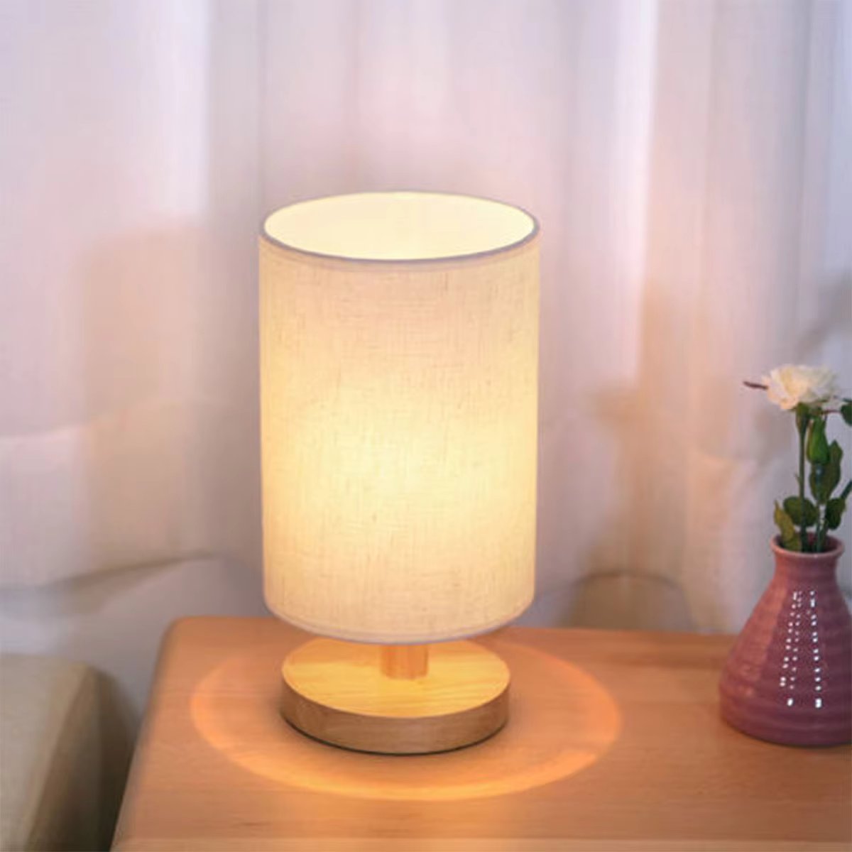 Table lamp with cylindrical white linen shade on round wooden base on bedside table beside pink ceramic vase with roses