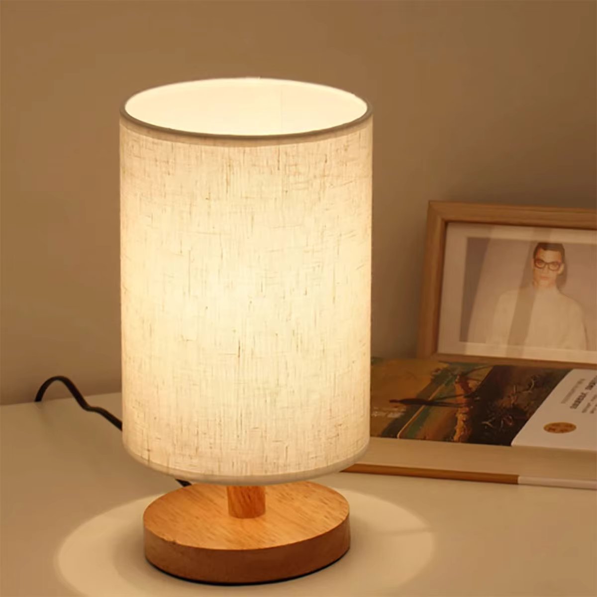 Table lamp with cylindrical linen shade and round wooden base on white desk beside framed photo and open magazine