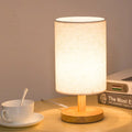 Table lamp with cylindrical white linen shade and round wooden base glowing on desk beside white cup and stacked books