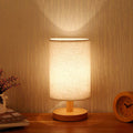 Table lamp with cylindrical linen shade on wooden base glowing against wood-effect wall beside wooden alarm clock and flowers