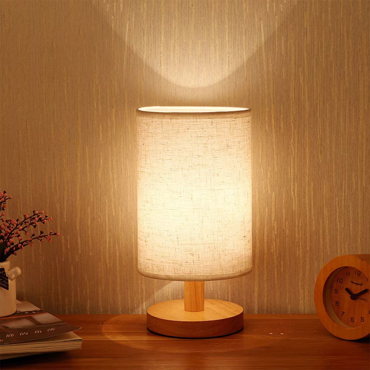 Table lamp with cylindrical linen shade on wooden base glowing against wood-effect wall beside wooden alarm clock and flowers