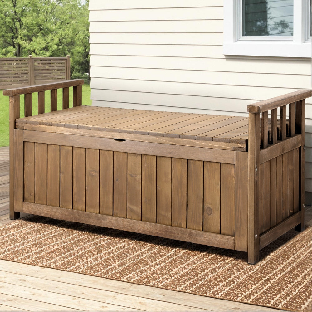 Brown timber outdoor storage bench on patio deck with woven rug, weatherboard house wall and trees in background