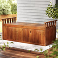 Timber outdoor storage bench on white rug, weatherboard house wall and green plants in background, closed lid
