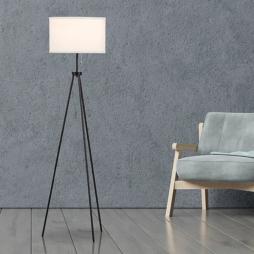 Tripod floor lamp with white cylindrical drum shade and three splayed black metal legs on white background