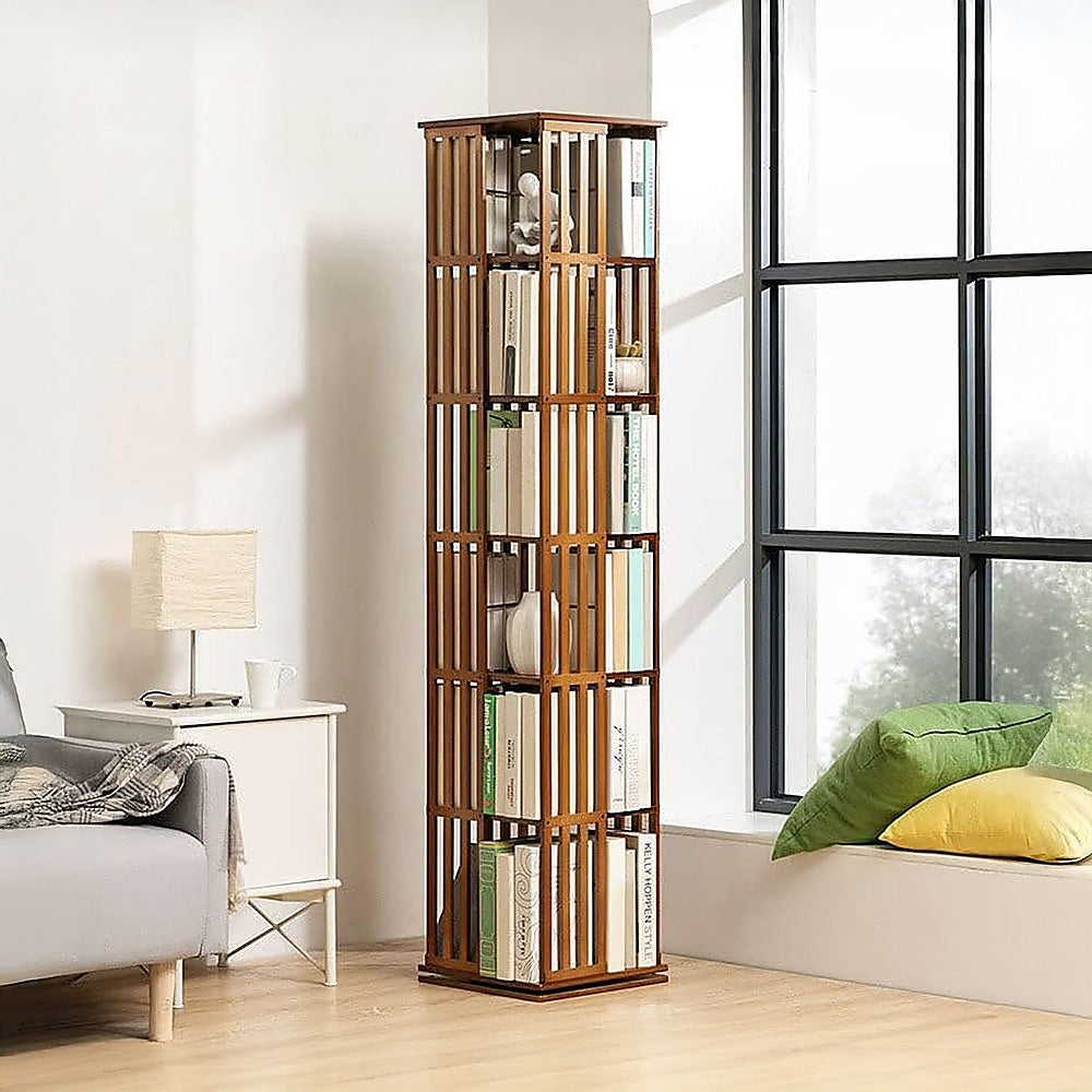 Walnut-finish slat rotating tower bookshelf filled with books in living room near large window, sofa and lamp visible