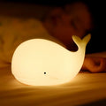 White whale-shaped silicone LED night light glowing warmly on surface beside sleeping person in dark room