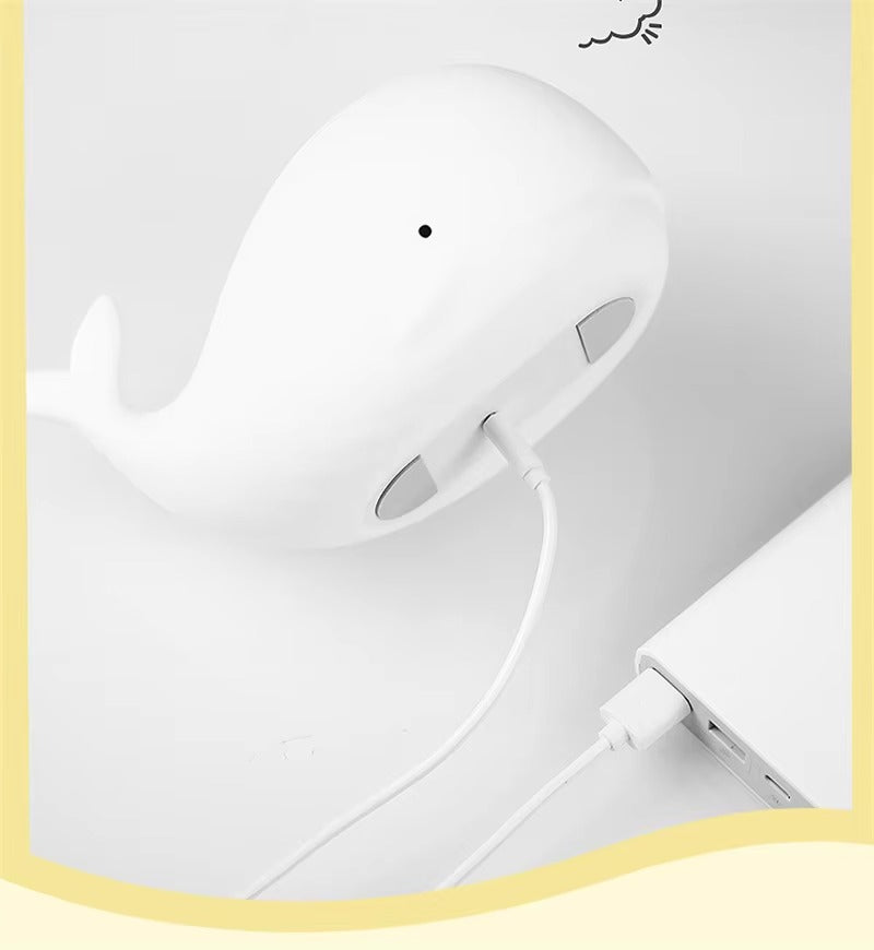 White whale-shaped silicone LED lamp being charged via USB cable connected to laptop port