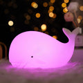 Whale-shaped silicone LED lamp glowing pink purple on fluffy surface with golden bokeh lights in dark background