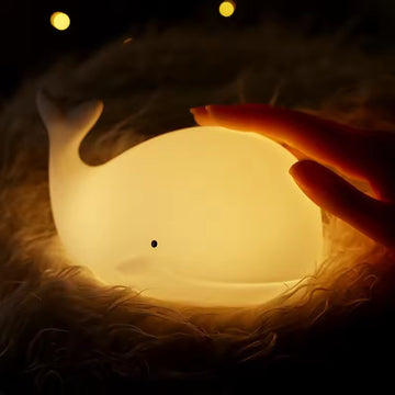 Glowing white whale-shaped silicone LED night light held in two hands on fluffy surface against dark background