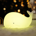 Whale-shaped silicone LED lamp glowing yellow green on fluffy surface with golden bokeh lights in dark background