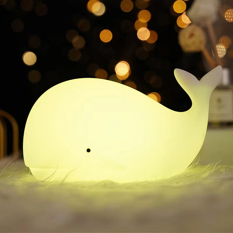 Whale-shaped silicone LED lamp glowing yellow green on fluffy surface with golden bokeh lights in dark background
