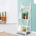 White 3-tier A-frame ladder shelf styled with books, aloe plant and hat boxes against teal wall with sideboard in background