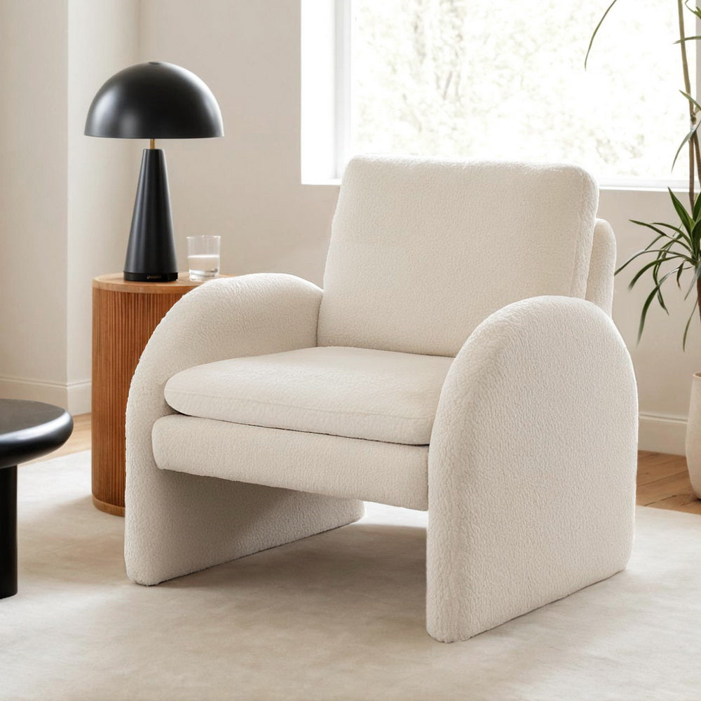 White boucle arch armchair in room with wooden side table, black dome lamp and glass, indoor plant, cream rug