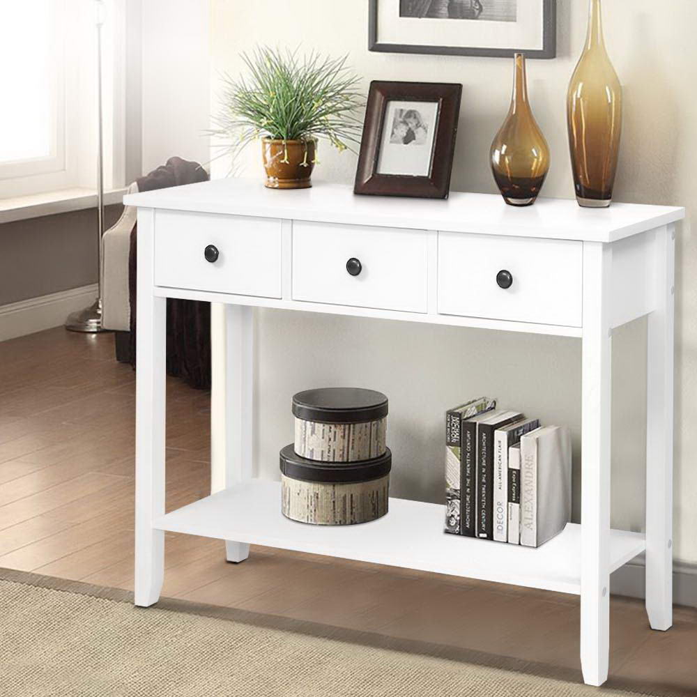 White console table with 3 drawers and bottom shelf in room; styled with plant, photo frame, amber vases, and books on shelf