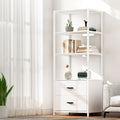White three-tier display shelf with two drawers styled with books and plants in a bright living room beside a window
