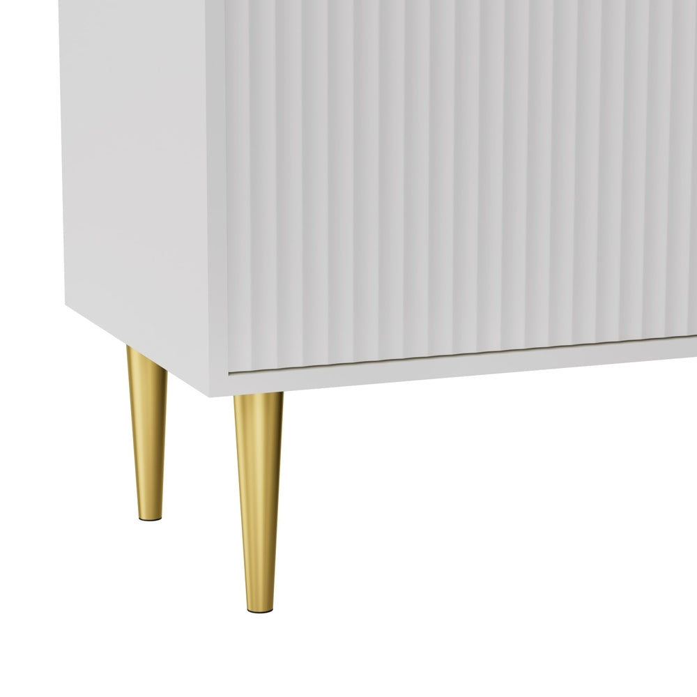 Close-up of white fluted TV unit lower corner showing 2 gold tapered metal legs against white background