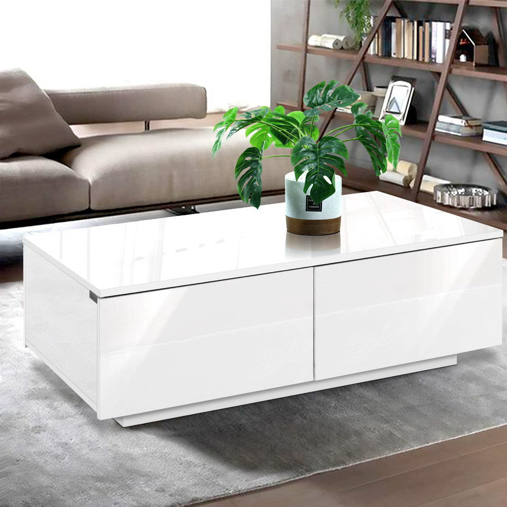 White gloss coffee table with two drawers on grey rug in living room with sofa and wooden bookshelf