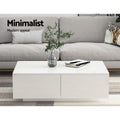 White gloss coffee table with two drawers in front of grey sofa, glass vase with plant and book on top
