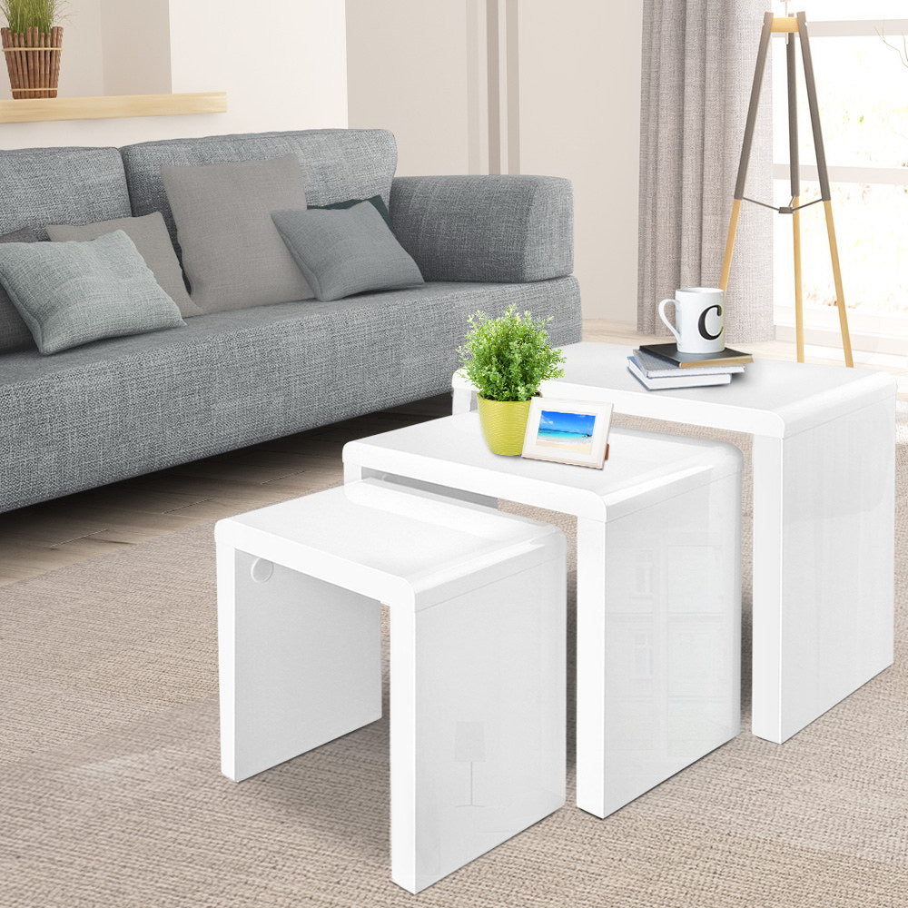 Set of three white high-gloss nesting tables in living room with grey sofa, plant and photo frame on top