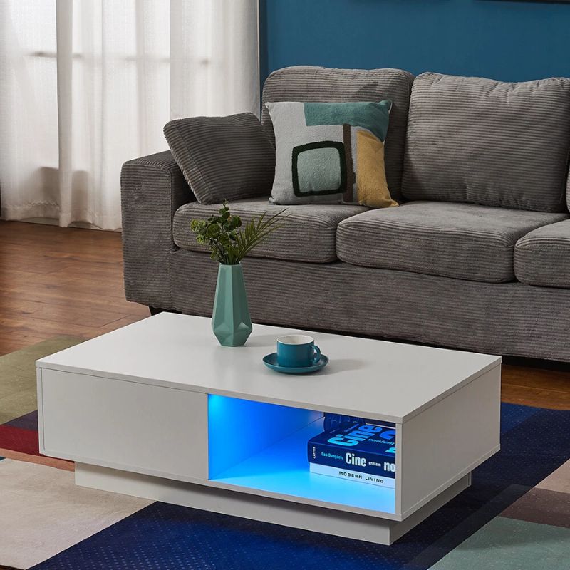 White high-gloss coffee table in living room, blue LED lit open shelf with books, teal vase and cup on top, grey sofa behind