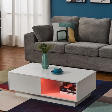White high-gloss coffee table in living room, open shelf with orange LED lighting and books, beside grey sofa and blue wall