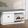 White shoe bench with grey fabric cushion left door closed and right flip-out compartment open showing shoes on parquet floor