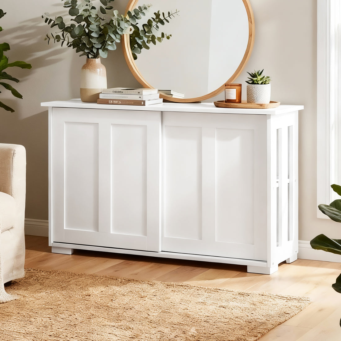 White sideboard with sliding doors in living room, top styled with books, eucalyptus vase, and round timber mirror on wall