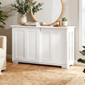White sideboard with sliding doors in living room, top styled with books, eucalyptus vase, and round timber mirror on wall
