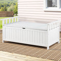 White timber outdoor storage bench on patio deck with woven rug, white weatherboard house wall and trees beyond