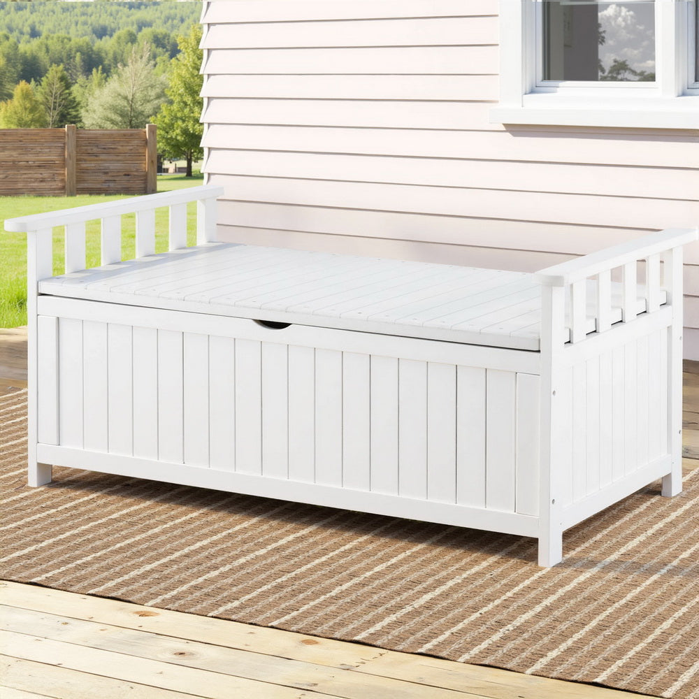 White timber outdoor storage bench on patio deck with woven rug, white weatherboard house wall and trees beyond