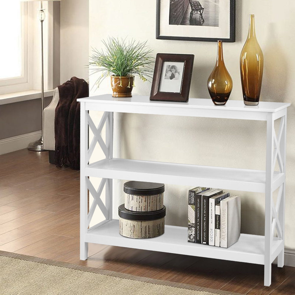 White console table with X-frame sides and two open shelves in room; styled with plant, photo frame, amber vases and books