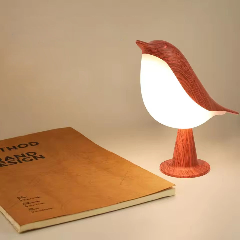 Red wood-grain bird table lamp glowing warm beside open Brand Design hardcover book, side angle on white surface